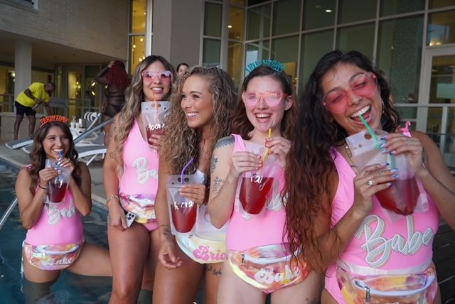Five women at a poolside bachelorette-style party in matching pink swimsuits and pastel fanny packs, laughing and sipping red drinks from clear pouch cups while wearing sunglasses.