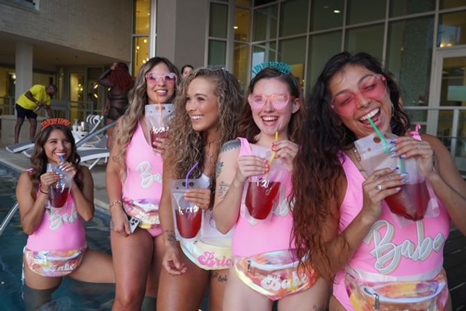 Five women at a poolside bachelorette-style party in matching pink swimsuits and pastel fanny packs, laughing and sipping red drinks from clear pouch cups while wearing sunglasses.