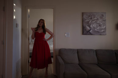 Woman in a red midi dress posing in a doorway of a cozy apartment living room, holding a pearl necklace next to a gray couch and rose wall art in soft, moody light.