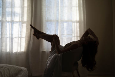 Silhouette of a woman lounging backward in a chair with legs raised, flowing dress and long hair against sunlit sheer curtains in a softly lit bedroom.