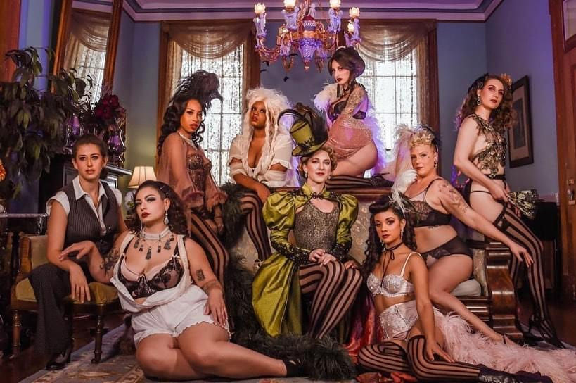 Nine performers in vintage burlesque and cabaret costumes — corsets, striped stockings, feathered headpieces and a top hat — posed in an ornate Victorian parlor with chandelier and draped windows.