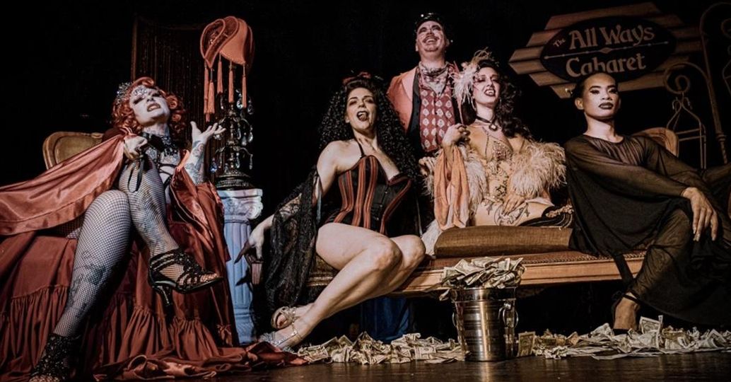 Five burlesque performers in vintage cabaret costumes pose on a dimly lit stage with corsets, feather boas, fishnets, scattered dollar bills and a metal tip bucket.