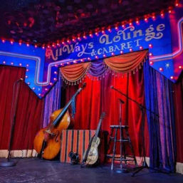 Intimate cabaret stage with red velvet curtains and blue marquee lights, an upright bass and acoustic guitar leaning by a striped box, microphones and a stool set for live music.