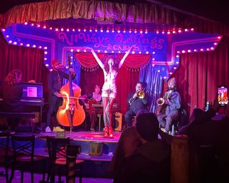 Burlesque performer striking a pose with arms raised on a small, colorful cabaret stage under marquee lights, backed by a live band (double bass, piano, trombone, saxophone) and an audience in an intimate nightclub setting.