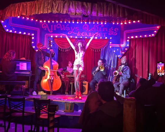 Burlesque performer striking a pose with arms raised on a small, colorful cabaret stage under marquee lights, backed by a live band (double bass, piano, trombone, saxophone) and an audience in an intimate nightclub setting.