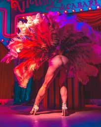 Burlesque performer on a cabaret stage, back to camera, posing with dramatic red and pink feather fans, rhinestone thong and glittering high-heeled showgirl boots under neon stage lights