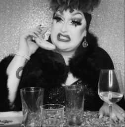 Black-and-white portrait of a glamorous drag performer in dramatic makeup and a feathered headpiece holding a banana to their ear like a phone, grimacing at a table with empty glasses and a wine glass against a sparkly backdrop.