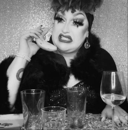 Black-and-white portrait of a glamorous drag performer in dramatic makeup and a feathered headpiece holding a banana to their ear like a phone, grimacing at a table with empty glasses and a wine glass against a sparkly backdrop.