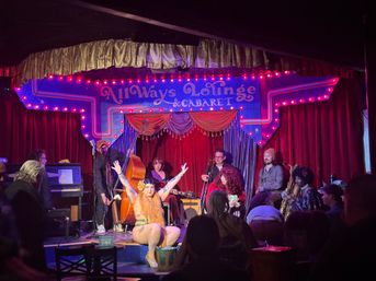 Intimate cabaret stage with red velvet curtains and neon marquee, a sparkly costumed performer with arms raised center-stage, live band (upright bass, piano, saxophone) behind and a seated audience in the foreground.