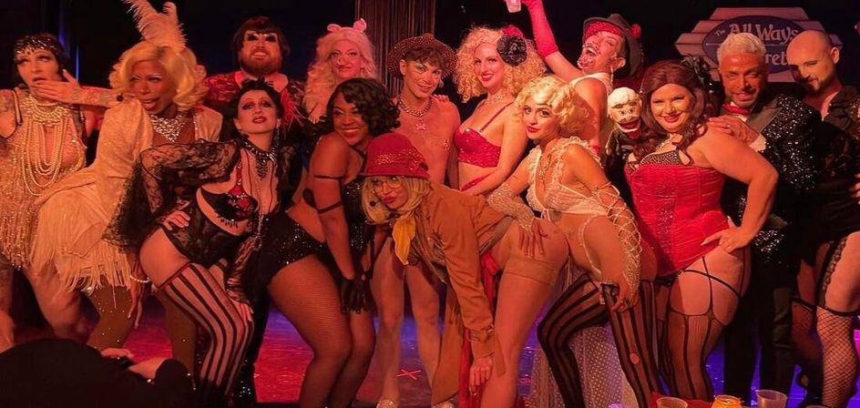 Group of cabaret and burlesque performers in vintage corsets, stockings and sequins posing together on a red-lit stage during a lively nightlife show