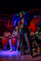 Performer in a rainbow sequin jumpsuit, blue faux-fur jacket and heart-shaped sunglasses striking a pose on a cabaret-style stage with red curtains and marquee lights, backed by percussionists on congas and a guitarist.