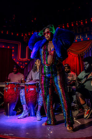 Performer in a rainbow sequin jumpsuit, blue faux-fur jacket and heart-shaped sunglasses striking a pose on a cabaret-style stage with red curtains and marquee lights, backed by percussionists on congas and a guitarist.