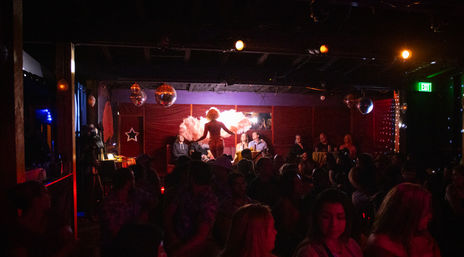Crowded dimly lit cabaret: backlit burlesque performer with large feather fans on a small stage under disco balls, audience seated and standing.