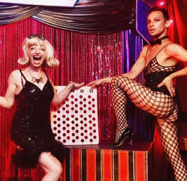 Two cabaret performers in sparkly black costumes and fishnet stockings pose on a glittery red stage beside an oversized Connect Four game prop