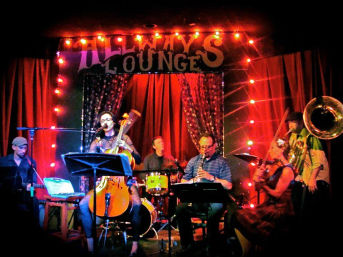 Live music on an intimate lounge stage: six-piece band under red curtains and string lights — singer with upright cello, drummer, clarinet player, violinist, tuba and keyboardist in warm colorful lighting.