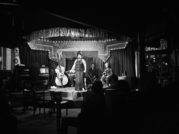 Black-and-white image of an intimate vintage cabaret stage: a glamorous singer in a sequined gown fronts a jazz combo with piano, upright bass, brass and saxophone while a seated audience watches.