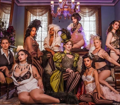 Nine performers in vintage burlesque and cabaret costumes — corsets, striped stockings, feathered headpieces and a top hat — posed in an ornate Victorian parlor with chandelier and draped windows.