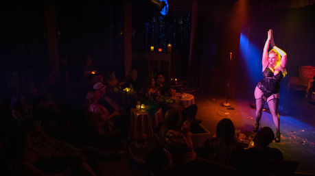 Burlesque performer in black lingerie strikes a pose on a small cabaret stage under colorful spotlights while an intimate, candlelit audience watches from tables.