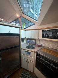 Compact yacht galley with stainless refrigerator, built-in microwave and stove, round sink on a laminate counter, and large angular skylight showing marina structure above