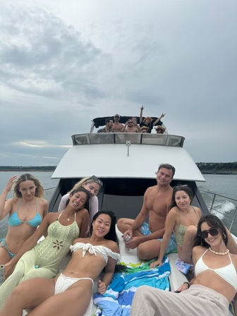 Cheerful group of friends lounging on the bow of a white yacht in coastal waters under a cloudy summer sky — boat day fun