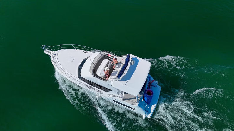 Aerial view of a white luxury motor yacht cruising through deep green water with passengers relaxing on the sunlit upper deck.