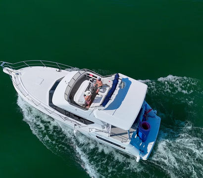 Aerial view of a white luxury motor yacht cruising through deep green water with passengers relaxing on the sunlit upper deck.
