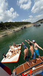 Young adults toasting with cups from a boat while friends lounge on inflatable platforms at a sunny lake cove with wooded shoreline and docks