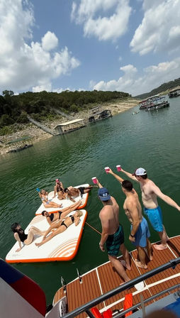 Young adults toasting with cups from a boat while friends lounge on inflatable platforms at a sunny lake cove with wooded shoreline and docks
