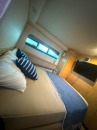 Cozy boat cabin interior with beige bed, blue striped pillows and fringed navy throw, rectangular porthole window and wall-mounted TV