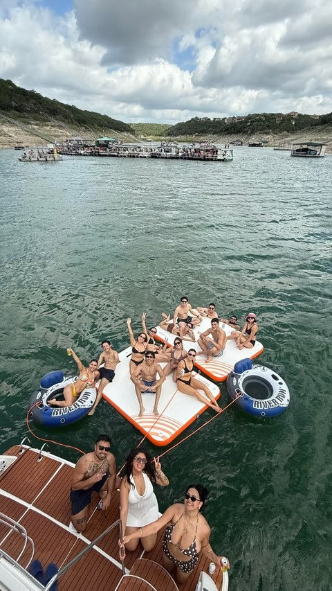 Group of people enjoying a summer boat party on an orange-and-white inflatable floating island with inner tubes on a scenic lake surrounded by green hills and a partly cloudy sky.