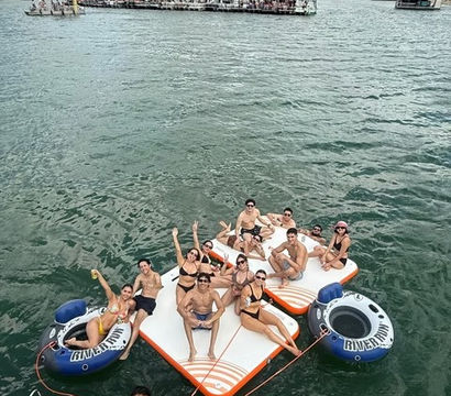 Group of people enjoying a summer boat party on an orange-and-white inflatable floating island with inner tubes on a scenic lake surrounded by green hills and a partly cloudy sky.
