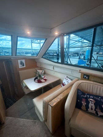 Cozy boat cabin dining nook with cream leather seating and table, patriotic pillows and centerpiece, large windows overlooking marina slips and covered dock.