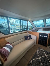 Yacht salon interior with cream leather sofa, striped and novelty pillows, built-in cabinet and large windows framing a marina with docked boats and blue sky.