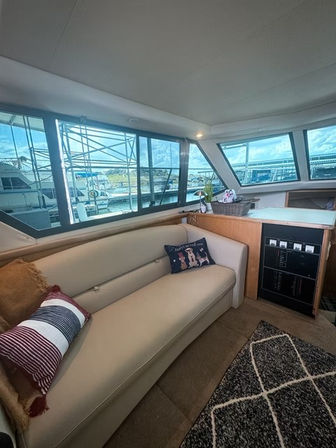 Yacht salon interior with cream leather sofa, striped and novelty pillows, built-in cabinet and large windows framing a marina with docked boats and blue sky.