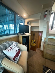 Cozy boat cabin interior at a marina dock: cream leather seat with a red-car throw pillow, small TV on a wooden cabinet, acoustic guitar in a nook, carpeted steps and marina slips visible through the window.