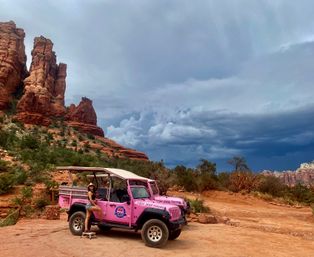 Bright pink open-top tour jeep parked on red sandstone ledge in Sedona, Arizona, framed by towering red rock formations, desert shrubs and a dramatic stormy blue-gray sky, a person posing beside the vehicle.