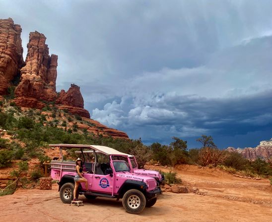 Bright pink open-top tour jeep parked on red sandstone ledge in Sedona, Arizona, framed by towering red rock formations, desert shrubs and a dramatic stormy blue-gray sky, a person posing beside the vehicle.