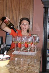 Person pouring white wine from a bottle into a row of stemmed tasting glasses on a marble bar counter in an indoor wine-tasting setting.