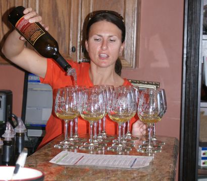 Person pouring white wine from a bottle into a row of stemmed tasting glasses on a marble bar counter in an indoor wine-tasting setting.