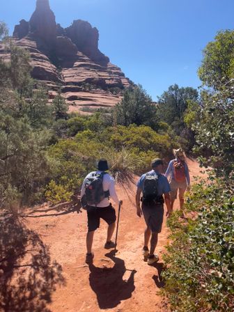 Three hikers with backpacks on a red-rock hiking trail through desert scrub toward layered sandstone cliffs under a clear blue Southwestern sky