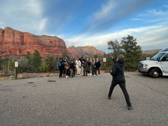 Group of people posing for a photo at sunset beside red rock cliffs in Sedona, Arizona, with a tour van parked nearby and a guide snapping the picture under a sweeping blue sky.