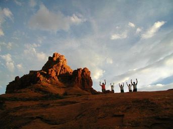 Silhouetted group of hikers raising arms on a sunlit red rock formation against a dramatic desert sky at sunset