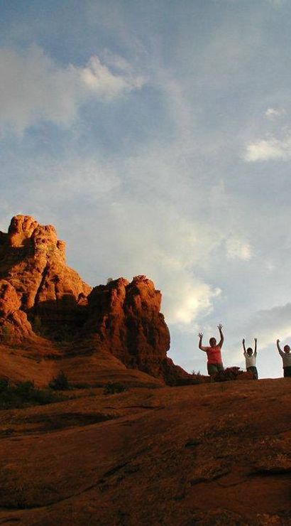 Silhouetted group of hikers raising arms on a sunlit red rock formation against a dramatic desert sky at sunset
