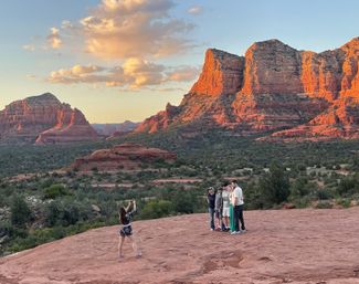 Person taking a photo of a small group on a red sandstone overlook at sunset with dramatic red-rock formations and desert scrub in Sedona, Arizona.