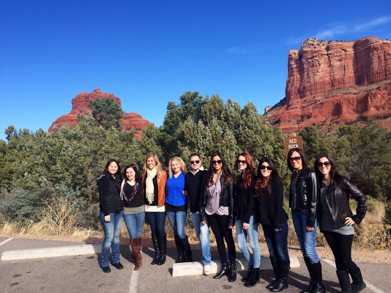 Group of people posing by a parking lot with towering red rock cliffs, juniper trees, and a 'Red Rock' sign under a clear blue sky in a southwestern desert landscape.