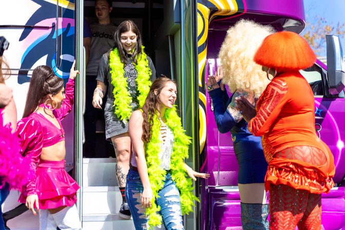 Group stepping off a bright purple party bus at an outdoor Pride celebration, wearing neon green feather boas and colorful drag costumes.