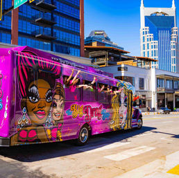 Vibrant pink drag-themed party bus with cartoon queen artwork and passengers waving from open windows on a sunny downtown street with modern skyscrapers
