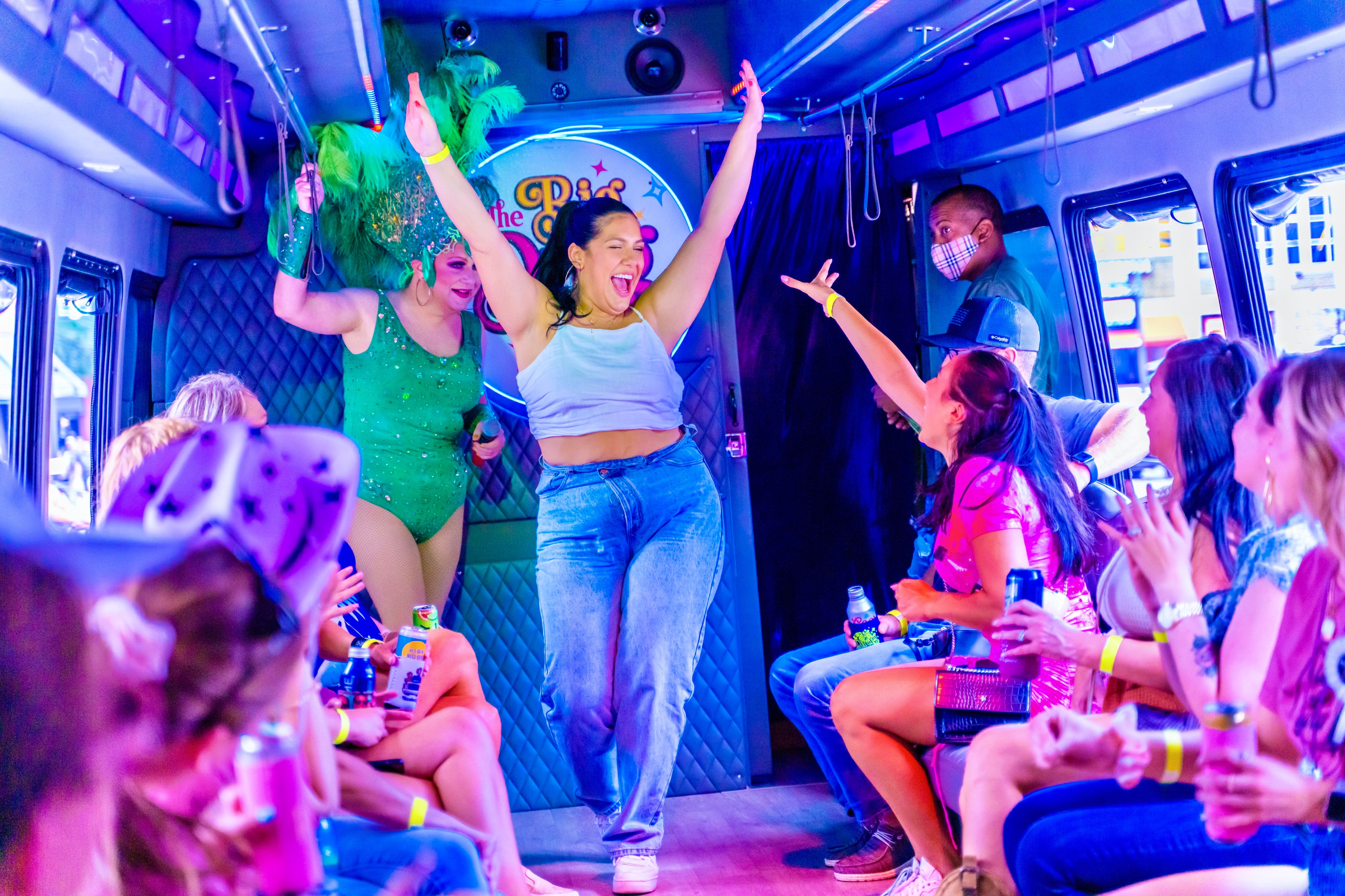 Neon-lit party bus interior with passengers cheering and holding drinks as a woman dances with arms raised while a drag performer in a green feather headdress entertains.