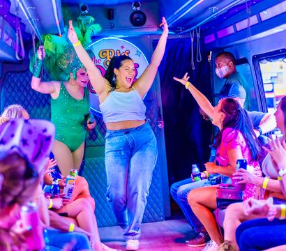 Neon-lit party bus interior with passengers cheering and holding drinks as a woman dances with arms raised while a drag performer in a green feather headdress entertains.