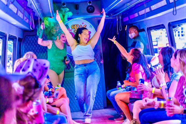 Neon-lit party bus interior with passengers cheering and holding drinks as a woman dances with arms raised while a drag performer in a green feather headdress entertains.
